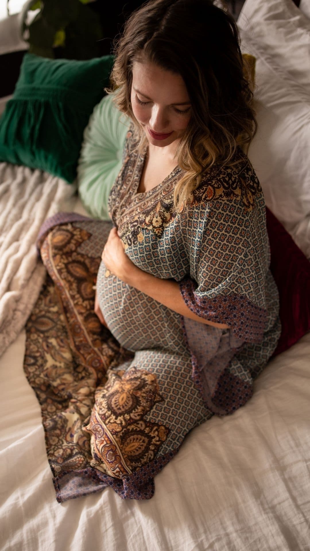 A pregnant woman seated on a bed, resting a hand on her belly